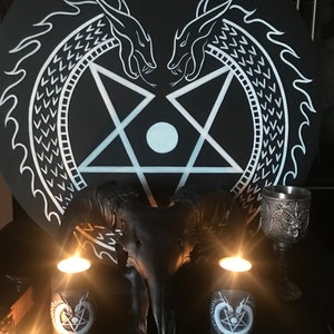 Puede incluir: Un pentagrama blanco y negro con dos dragones blancos enfrentados. El pentagrama está rodeado por un círculo con un patrón blanco. Un cráneo de carnero negro se encuentra frente al pentagrama. Tres velas negras con diseños blancos están sobre la mesa. Un cáliz de plata está sobre la mesa.