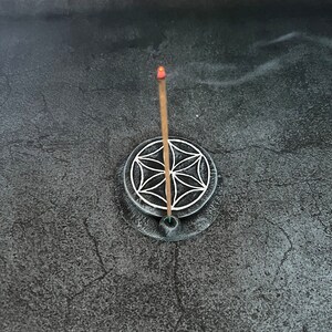 May include: A black incense holder with a white geometric design and a burning incense stick.