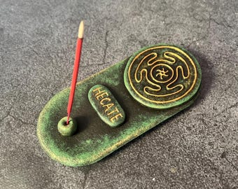 May include: A green ceramic incense holder with a weathered finish. The holder features a round incense burner with a spiral design, a small rectangular piece with the word "HECATE", and a hole for an incense stick. A red incense stick is inserted.
