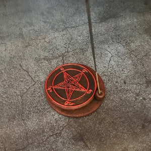 May include: A black and red ceramic incense burner with a pentagram design. A black incense stick is placed in the burner.