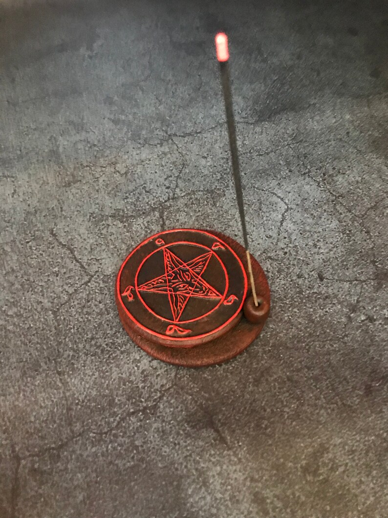 May include: A black and red ceramic incense burner with a pentagram design. A black incense stick is placed in the burner.