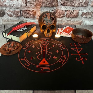 May include: A black altar cloth with red sigils and symbols, a skull with a pentagram on its forehead, two white candles, a small bowl, a deck of cards, and a book with a red feather on top.