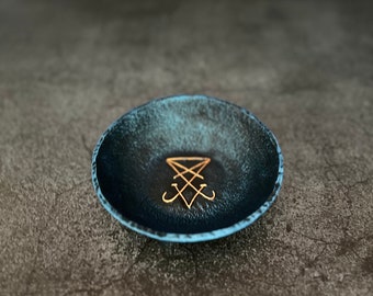 May include: A dark blue ceramic dish with a gold sigil in the center. The dish is sitting on a gray textured surface.