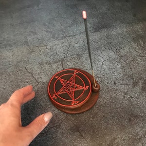 May include: A brown ceramic incense holder with a red pentagram design. A black incense stick is burning in the holder.