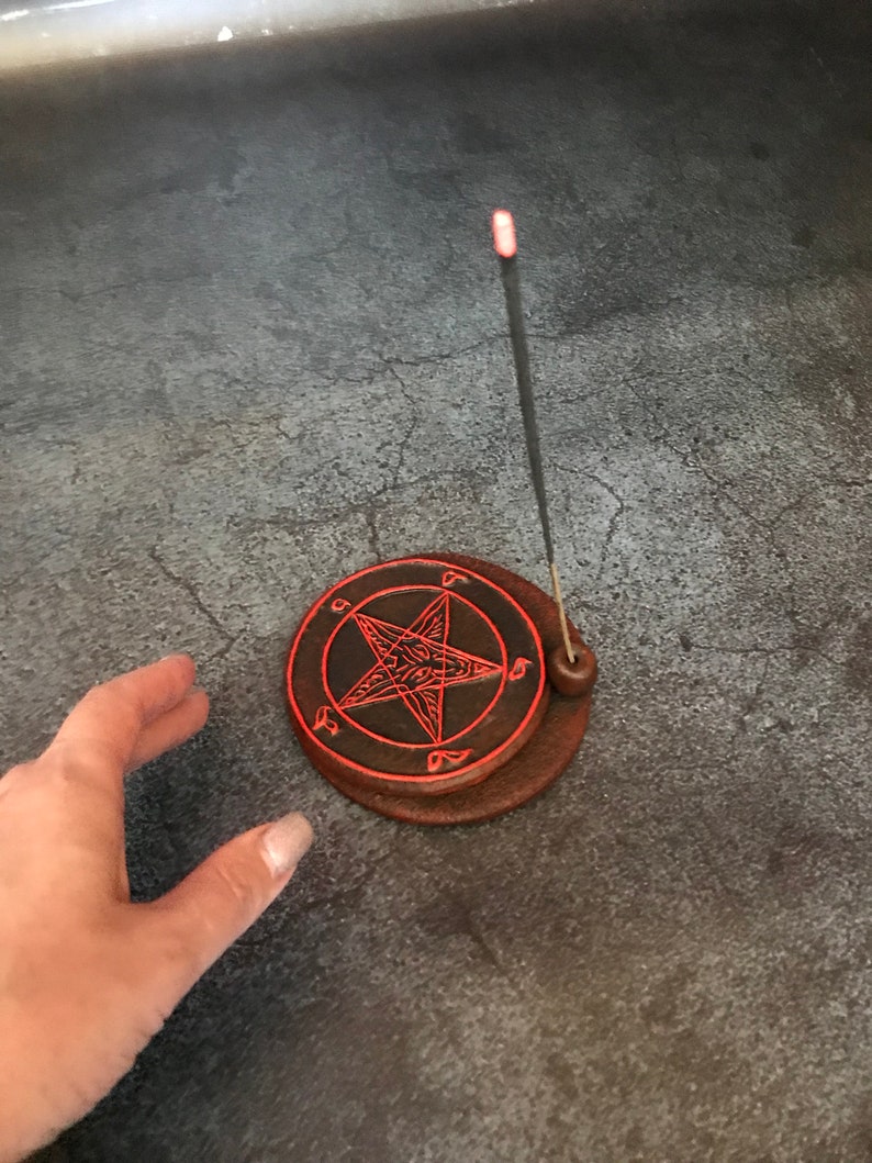 May include: A brown ceramic incense holder with a red pentagram design. A black incense stick is burning in the holder.