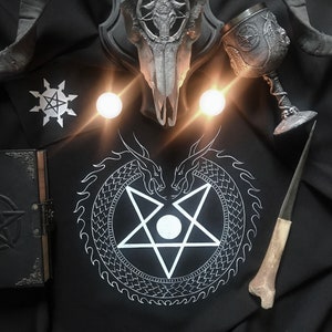 Puede incluir: Un fondo de tela negro con un pentagrama blanco rodeado de un diseño de serpiente. Un cráneo de animal plateado con cuernos está montado en una placa de madera. Un cáliz plateado con un diseño de pentagrama se encuentra sobre la tela. Un hueso y un pequeño libro negro también están sobre la tela.