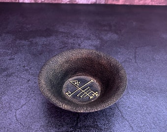 May include: A small, black, round bowl with a gold-colored symbol in the center. The bowl is made of metal and has a textured surface.
