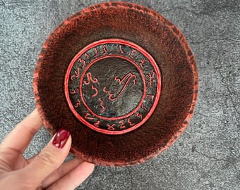 May include: A red and black ceramic dish with a circular design featuring a symbol and runes. The dish is approximately 5 inches in diameter.