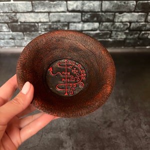 Peut inclure: Un petit bol rond brun foncé avec une surface texturée et un bord rouge. L'intérieur du bol présente une base noire avec un symbole rouge. Le bol est tenu par une personne.