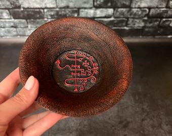 May include: A small, round, dark brown bowl with a textured surface and a red rim. The bowl's interior features a black base with a red symbol. The bowl is held by a person.