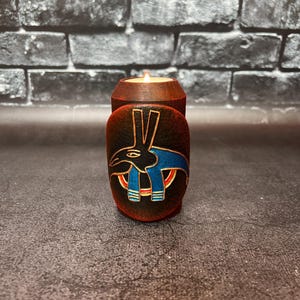 以下が含まれることがあります： 点灯したろうそくが入った木製のキャンドルホルダー。ホルダーは濃い茶色の仕上げで、黒、青、赤の様式化された鳥のデザインが施された装飾的な楕円形のパネルが付いています。背景は暗いレンガの壁です。