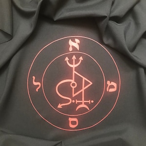 Pode incluir: Um selo vermelho sobre um fundo de tecido preto. O selo é um círculo com um tridente, um triângulo e uma lua crescente dentro. As letras hebraicas "Nun", "Shin", "Mem" e "Dalet" são escritas ao redor do círculo.