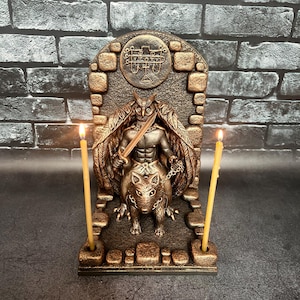 Könnte beinhalten: Eine bronzefarbene Statue einer geflügelten Figur, die ein Schwert hält und auf einem wolfsähnlichen Wesen reitet. Die Statue hat ein detailliertes Design mit einem eulenartigen Gesicht und wird von zwei brennenden Kerzen flankiert. Der Hintergrund ist eine Backsteinmauer.