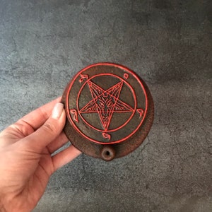 May include: A dark brown ceramic incense burner with a red pentagram design. The burner has a small hole in the center for incense sticks.