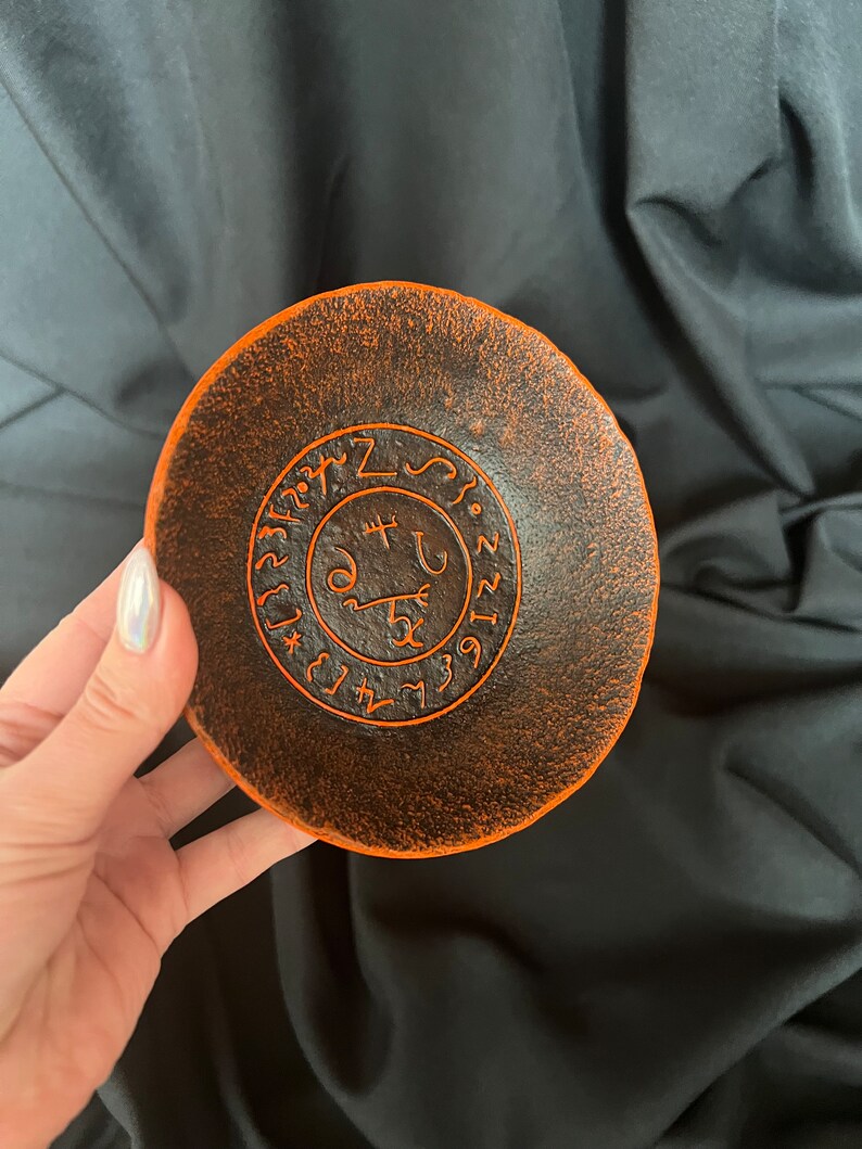 K&ouml;nnte beinhalten: Orangefarbene und schwarze Keramikschale mit einem kreisf&ouml;rmigen Design, das ein Symbol und Zahlen zeigt. Das Symbol ist von einem Kreis mit Zahlen umgeben.