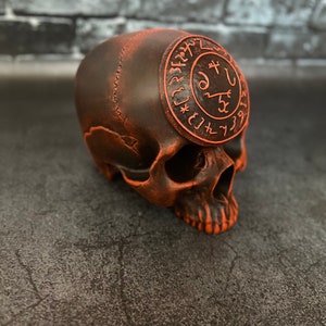 Op de afbeelding: Een gedetailleerd, donkerbruin schedelbeeldje met een oranje, cirkelvormig embleem bovenop, met ingewikkelde symbolen. De schedel heeft een verweerde, getextureerde uitstraling, met oranje accenten. Donkergrijze achtergrond.