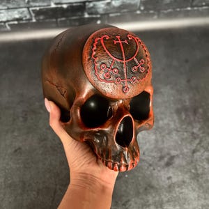 Op de afbeelding: Een donkerbruine schedel met een rood zegel op het voorhoofd. Het zegel is een cirkel met een kruis en andere symbolen erin. De schedel is gemaakt van hars en is een decoratief object.