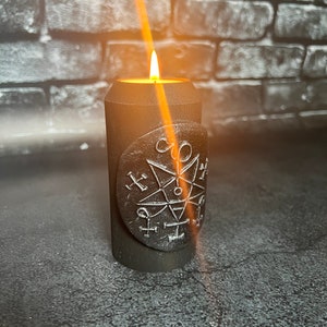 Pode incluir: Uma vela preta com um pentagrama e outros símbolos gravados no lado. A vela está acesa e a chama projeta um brilho quente.
