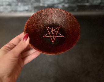 May include: A small, dark brown bowl with a red pentagram symbol inside. The bowl is made of ceramic and has a rough, textured surface.
