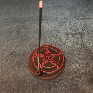 May include: A black incense stick with a red tip is burning on a brown ceramic incense holder with a red pentagram design.