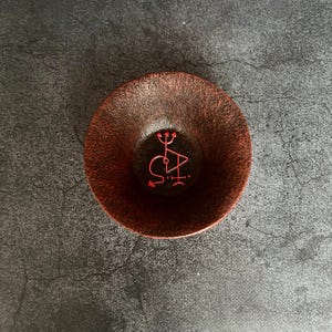 Pode incluir: Uma tigela pequena, de cor vermelho escuro, com uma borda elevada. A tigela tem um símbolo vermelho desenhado no interior, que se assemelha a um tridente com um círculo e um triângulo.
