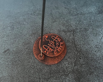 May include: A round, copper-colored metal incense holder with a textured finish and a raised circular center featuring an engraved sigil. A single black incense stick with a glowing red tip is inserted into the small hole on the base.
