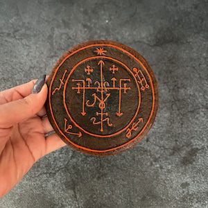 May include: A circular, brown, clay sigil with orange painted symbols and lines. The sigil is surrounded by a circle with an arrow pointing to the right.