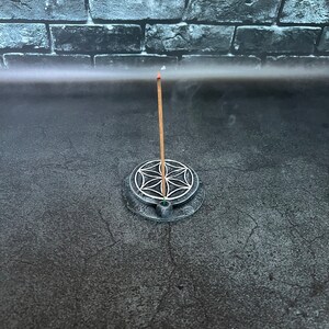 May include: A black incense holder with a circular design featuring a flower of life symbol. A wooden incense stick is burning in the holder, with smoke rising from the top.
