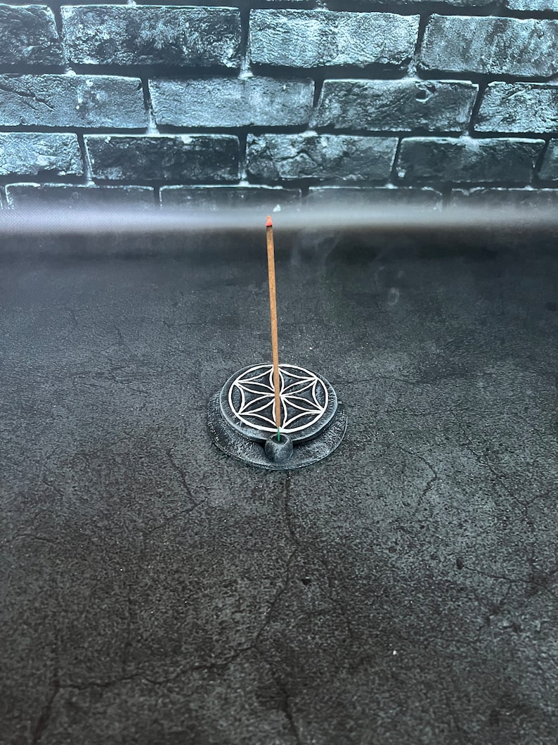 May include: A black incense holder with a circular design featuring a flower of life symbol. A wooden incense stick is burning in the holder, with smoke rising from the top.