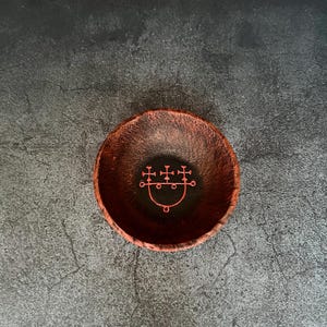 Könnte beinhalten: Eine kleine, runde Schale aus Terrakotta mit schwarzem Innenraum. Ein rotes Sigil ist auf der Innenseite der Schale gemalt.