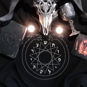 Könnte beinhalten: Ein schwarzer Stoff mit einem weißen Kreis, der einen Stern mit hebräischen Buchstaben enthält. Es gibt einen Schädel mit Hörnern, einen silbernen Kelch, ein schwarzes Buch mit einem Pentagramm und eine Tarotkarte mit einem Dämon darauf.