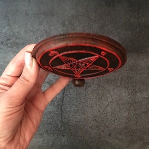 May include: A wooden round coaster with a red pentagram design. The pentagram has a goat's head in the center. The coaster has a small knob on the bottom.