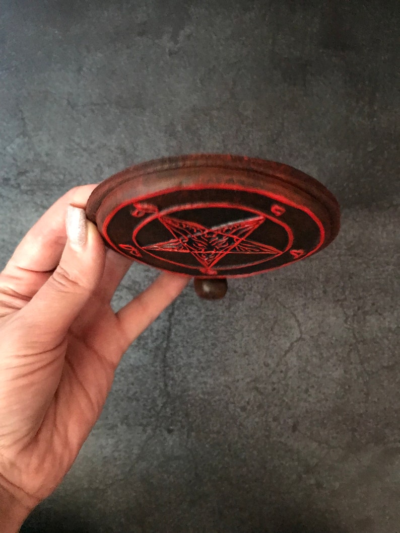 May include: A wooden round coaster with a red pentagram design. The pentagram has a goat's head in the center. The coaster has a small knob on the bottom.
