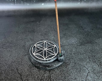 Op de afbeelding: Een zwarte keramische wierookhouder met een geometrisch ontwerp en een brandende wierookstok. De houder staat op een grijs oppervlak.