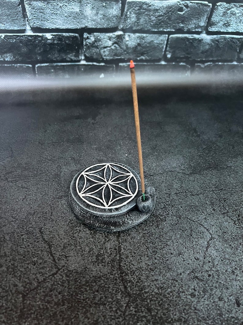 May include: A black ceramic incense holder with a geometric design and a single incense stick burning. The holder is on a gray surface.