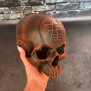 May include: A dark brown skull with an orange painted sigil on the forehead. The skull is made of resin and has a detailed design.