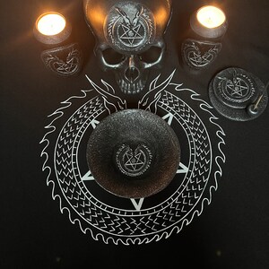 Op de afbeelding: Een zwarte schedel met een pentagram-ontwerp op het voorhoofd zit op een zwarte altaar doek met een wit pentagram-ontwerp. Twee zwarte kaarsenhouders met pentagram-ontwerpen en brandende kaarsen bevinden zich aan weerszijden van de schedel. Een zwarte wierookbrander met een pentagram-ontwerp bevindt zich in de rechterbenedenhoek van de afbeelding.