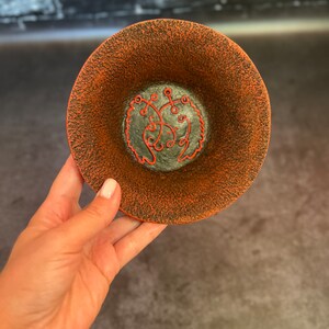 Puede incluir: Un pequeño plato redondo de cerámica naranja con un diseño negro y rojo en el centro. El plato tiene una superficie texturizada.
