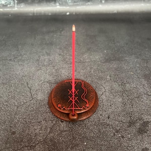 May include: A red incense stick is lit and placed in a brown, circular incense holder. The holder has a carved design in red and a small wooden ball at the base. The background is a dark gray surface.