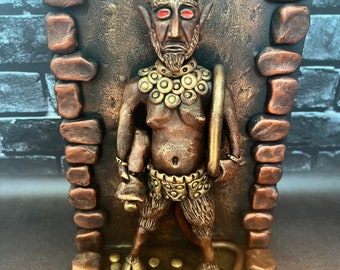 Può includere: Una scultura color bronzo di una figura cornuta con occhi rossi, barba e coda, in piedi davanti a una parete con texture in pietra. La figura indossa un collare circolare e un perizoma, tiene in mano un oggetto circolare, con monete d'oro e un sigillo ai piedi.