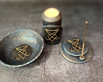 May include: A black wooden candle holder with a gold sigil, a black metal incense burner with a gold sigil, and a black metal bowl with a gold sigil. The candle holder has a small white candle inside.