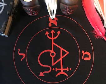 May include: A black cloth with a red circle and a sigil inside. The sigil is a triangle with a circle inside and a trident above it. There are Hebrew letters around the circle. Two candles are on the cloth, and a white animal skull is in the center.