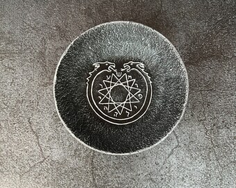 May include: A round, black coaster with a textured surface. The coaster features a white, stylized illustration of two dragons encircling a star-shaped design with symbols. The coaster is likely made of stone or a similar material.
