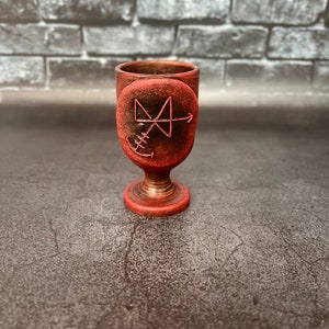 Może przedstawiać: Czerwono-brązowy kielich ceramiczny z okrągłym wzorem przedstawiającym czerwony symbol i kotwicę. Kielich stoi na szarym tle.
