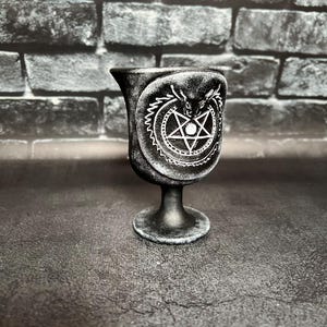 Puede incluir: Copa negra con un pentagrama blanco y un diseño de criatura alada blanca. La copa tiene una superficie texturizada y una base redonda.