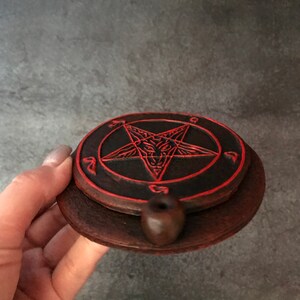 May include: A black and red ceramic incense burner with a pentagram design. The burner has a small hole in the center for incense sticks.