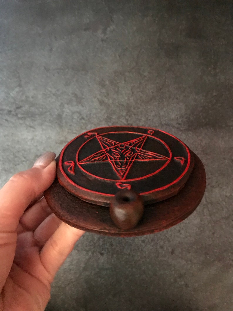 May include: A black and red ceramic incense burner with a pentagram design. The burner has a small hole in the center for incense sticks.
