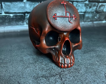 May include: A dark reddish-brown skull sculpture featuring a circular emblem on the forehead. The emblem displays a red linear symbol with cross-like terminals. The skull has deep-set eye sockets and a detailed jawline with visible teeth.