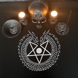 Könnte beinhalten: Ein schwarzer Altartuch mit einem weißen Pentagramm, das von einem Schlangendesign umgeben ist. Es gibt zwei schwarze Kerzenhalter mit brennenden Kerzen, einen schwarzen Schädel und einen schwarzen Räucherstäbchenhalter mit einem brennenden Räucherstäbchen.