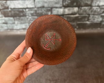 Op de afbeelding: Een klein, rond, bruin keramiek schaaltje met een rood ontwerp van een symbool met een pijl en een anker. Het schaaltje wordt vastgehouden door een hand.